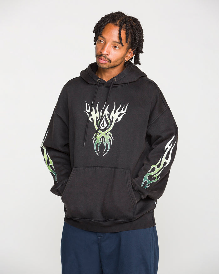 A person wearing the Volcom Men's Stone Trip Pullover—a washed-black hoodie featuring white and green flame designs on the chest and sleeves—stands with hands in the front pocket, looking slightly to the side.