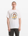 A man with short hair wears the Volcom Men's Stoned In Paradise Short Sleeve Tee, featuring "Stoned in Paradise" text and parrot graphic, paired with dark shorts, standing against a plain white background.