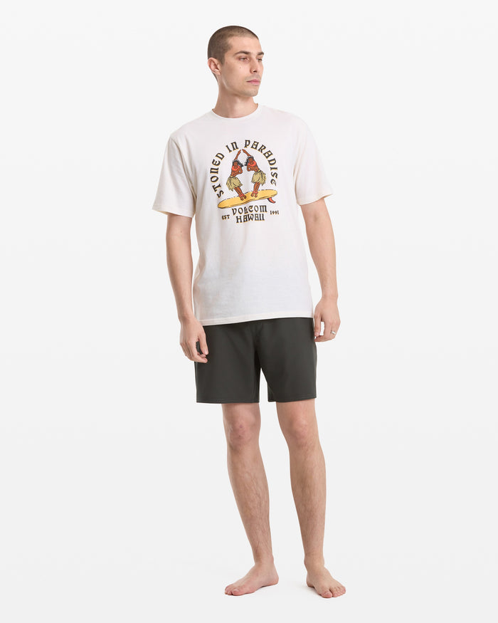 A barefoot man stands against a white background, wearing the Volcom Men's Stoned In Paradise Short Sleeve Tee with a colorful graphic and black shorts. He looks slightly to the side, arms relaxed by his sides.