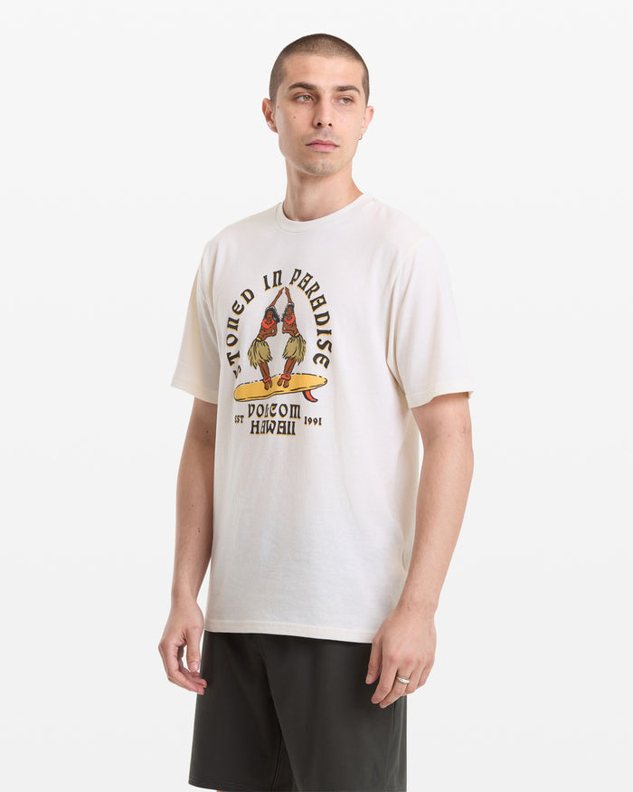 A person wears the Volcom Men's Stoned In Paradise Short Sleeve Tee with a graphic and dark shorts, standing against a plain white background.