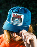 A person with long hair and sunglasses adjusts the brim of the Volcom Men's Stoney Mountain Hi Adjustable Hat, featuring an aqua blue cotton twill build and a branded patch, set against a dark, blurred background.