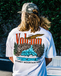 A person with long hair wears a camouflage cap and the Volcom Men's Stoney Mountain Short Sleeve Tee, made from 100% cotton with a vibrant waterfall, mountains, eagle, and trees graphic, plus VOLCANO and STONEY MOUNTAIN HIGH on the back.