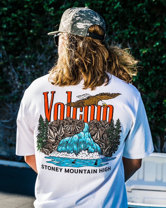 A person with long hair wears a camouflage cap and the Volcom Men's Stoney Mountain Short Sleeve Tee, made from 100% cotton with a vibrant waterfall, mountains, eagle, and trees graphic, plus VOLCANO and STONEY MOUNTAIN HIGH on the back.