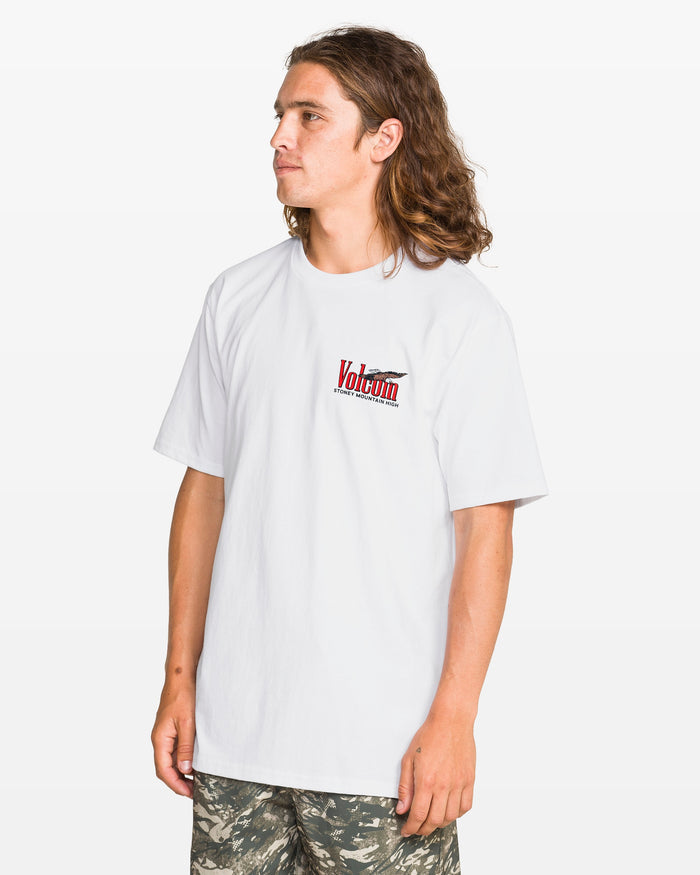 A person with long brown hair wears the Volcom Men's Stoney Mountain Short Sleeve Tee in white, featuring a small chest logo, paired with camo shorts, standing against a plain white background.