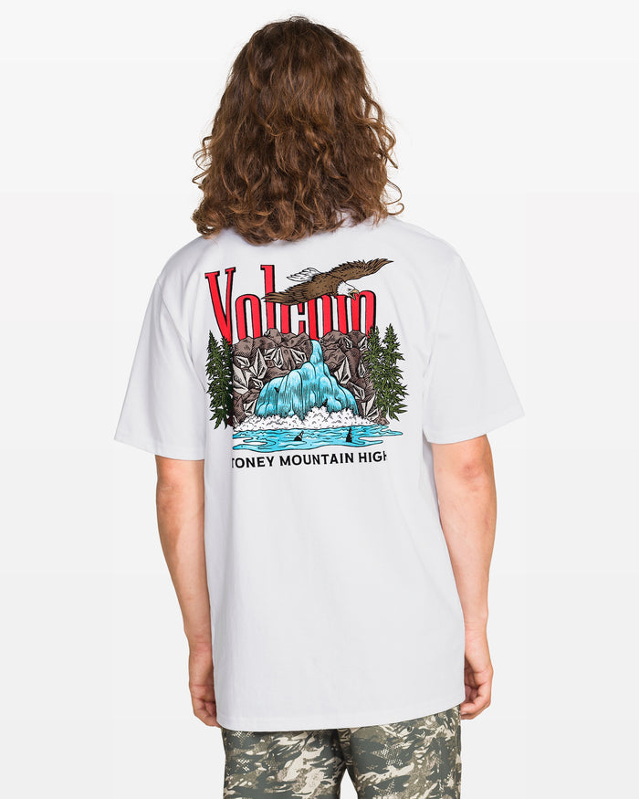 A person with long hair wears the Volcom Men's Stoney Mountain Short Sleeve Tee, a classic fit 100% cotton shirt with a colorful mountain and waterfall graphic on the back, paired with camouflage shorts.