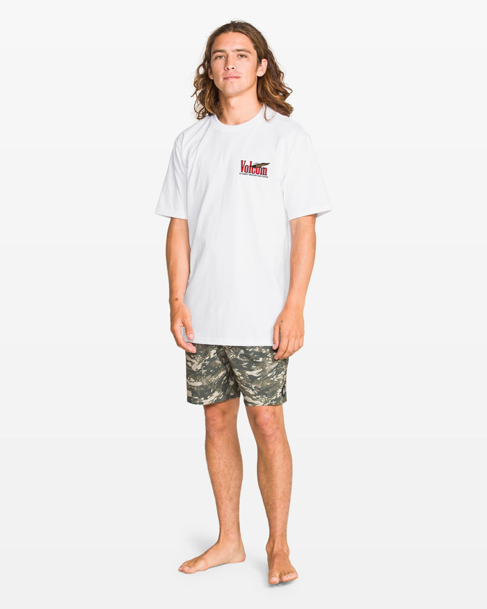 A person with long hair stands barefoot on a white background, wearing the Volcom Men's Stoney Mountain Short Sleeve Tee, a classic fit 100% cotton tee with a red and black chest logo, paired with camouflage shorts.