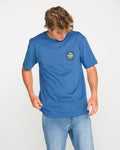A person dressed in jeans and a blue, classic fit Volcom Men's Stoneyguy Short Sleeve Tee looks down while holding the shirt’s hem. Made from premium cotton, the t-shirt features a small, round soft-hand screen print graphic on the left chest. The background is plain white.