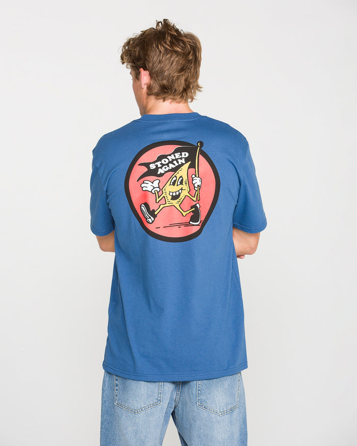 A person wearing the Volcom Men's Stoneyguy Short Sleeve Tee in blue and light jeans is seen from behind. The shirt features a soft-hand screen print of a cartoon slice of pizza holding a cane, with the text "STONED AGAIN" above it in a red circle.