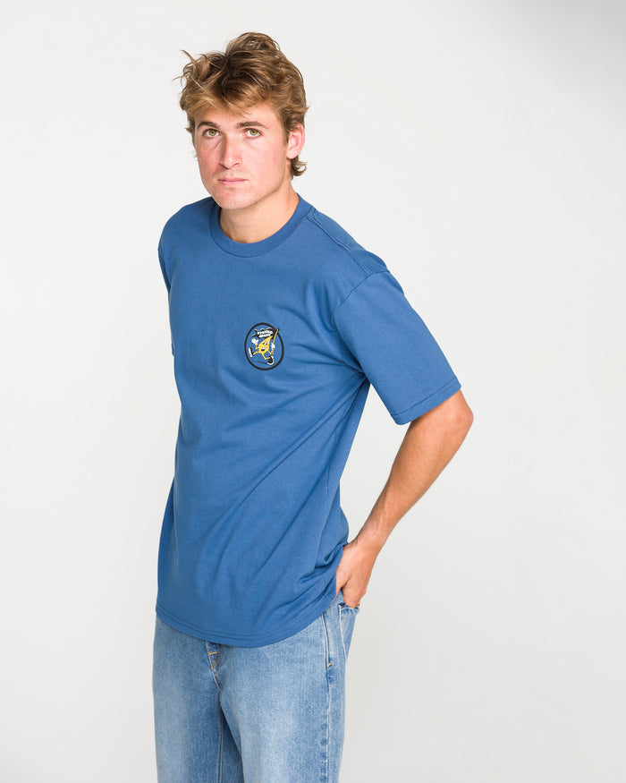 A young man with light brown hair wears the Volcom Men's Stoneyguy Short Sleeve Tee in blue, which features a small graphic on the chest rendered in a soft-hand screen print. He pairs it with light blue jeans and stands against a plain white background, looking at the camera with a neutral expression.