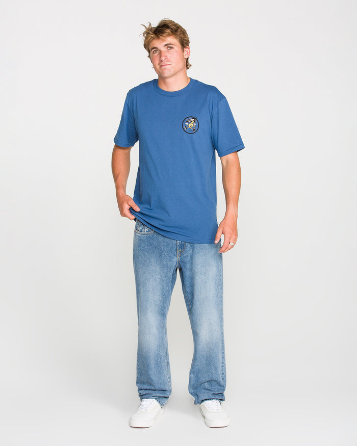 A young man wearing the Volcom Men's Stoneyguy Short Sleeve Tee, featuring a soft-hand screen print graphic on the chest, paired with light blue jeans and white sneakers, stands against a plain white background.