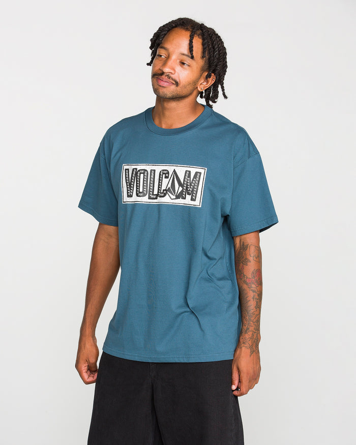 A man with tattoos wears the Volcom Men’s Studded Short Sleeve Tee in blue, crafted from premium ringspun cotton, paired with black shorts. He stands against a plain white background with short dreadlocks and looks slightly to the side, displaying a relaxed expression.