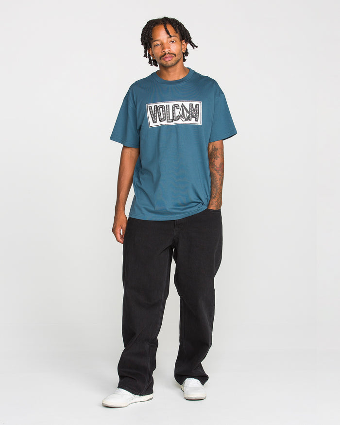 A man with short dreadlocks and a tattoo on his left forearm stands against a white background wearing the Volcom Men’s Studded Short Sleeve Tee in teal, made from premium ringspun cotton, paired with loose black pants and white shoes.