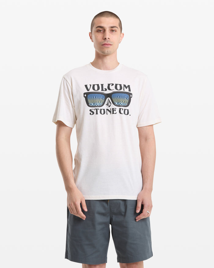 A man with short hair wears a white Volcom Men's Sunnies Short Sleeve Tee featuring a sunglasses graphic and dark gray shorts, standing against a plain white background.