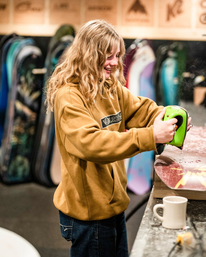 Wearing the Volcom Men’s Super Stoned Pullover, a person with long blonde hair smiles while brushing wax onto a snowboard with an iron in a shop. Several snowboards can be seen in the background, and a white mug sits nearby on the table. The brushed-back fleece hoodie from Volcom adds extra comfort to their look.