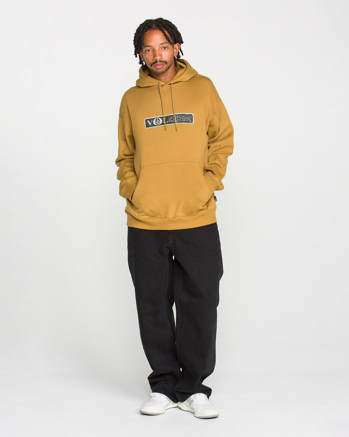A person stands against a plain white background, facing forward with hands in the pocket of a tan Volcom Men's Super Stoned Pullover hoodie made from recycled polyester and brushed-back fleece, paired with black pants and white sneakers.