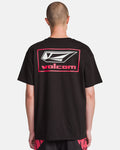 A person with a shaved head, seen from behind, wears a black Volcom Men's Surf Vitals Modstone Short Sleeve Tee with a large white and red logo and text on the back, paired with black and red shorts.