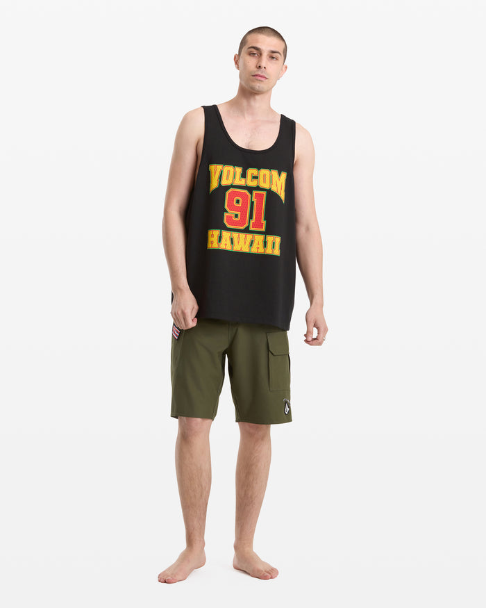 A man stands barefoot against a white background, wearing the Volcom Men's Swish Tank—an oversized black tank top with red and yellow text—and olive green cargo shorts.
