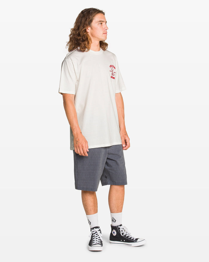 A young man with long hair stands sideways against a plain white background, wearing a Volcom Men's Temptations Short Sleeve Tee in white, paired with checkered gray shorts, white socks, and black Converse sneakers.