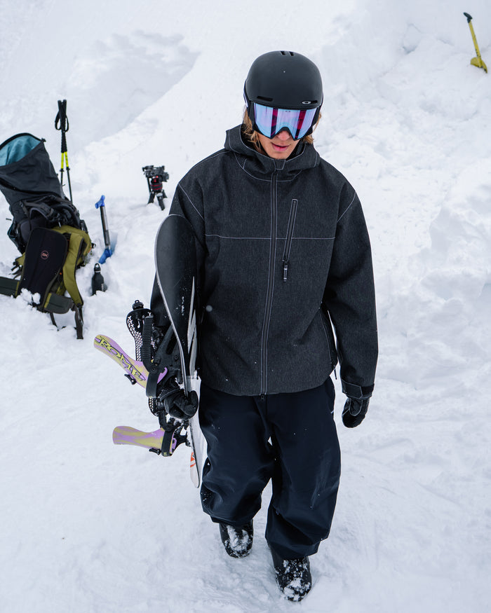 Clad in Volcom’s Men’s Top Stitch Jacket—featuring a baggy fit and 10K waterproof protection—a person in black winter gear and goggles stands in the snow, holding skis and poles. Backpacks and equipment appear in the background.