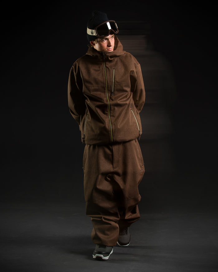 A person wearing the Volcom Men’s Top Stitch Jacket in a brown, baggy fit with 10K/10K protection, along with matching baggy brown pants, a black beanie, goggles, and sneakers stands against a dark background with their hands behind their back.