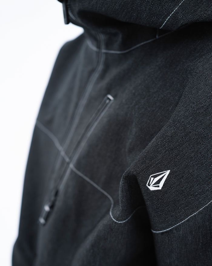 Close-up of the Volcom Men’s Top Stitch Jacket in black, showcasing a baggy fit, distinctive white geometric logo on the sleeve, bold white stitching, and a partially visible zipper against a softly blurred light background. With 10K waterproofing, this jacket keeps you dry while elevating your style.
