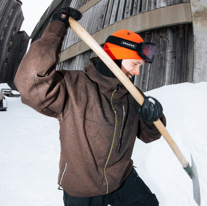A person shovels snow outside next to a wooden building, wearing the Volcom Men’s Top Stitch Jacket in a baggy fit brown style with PFAS-free DWR and 10K/10K protection, paired with an orange hat and goggles. The image, taken from a low angle, captures them mid-action in this winter-ready gear.