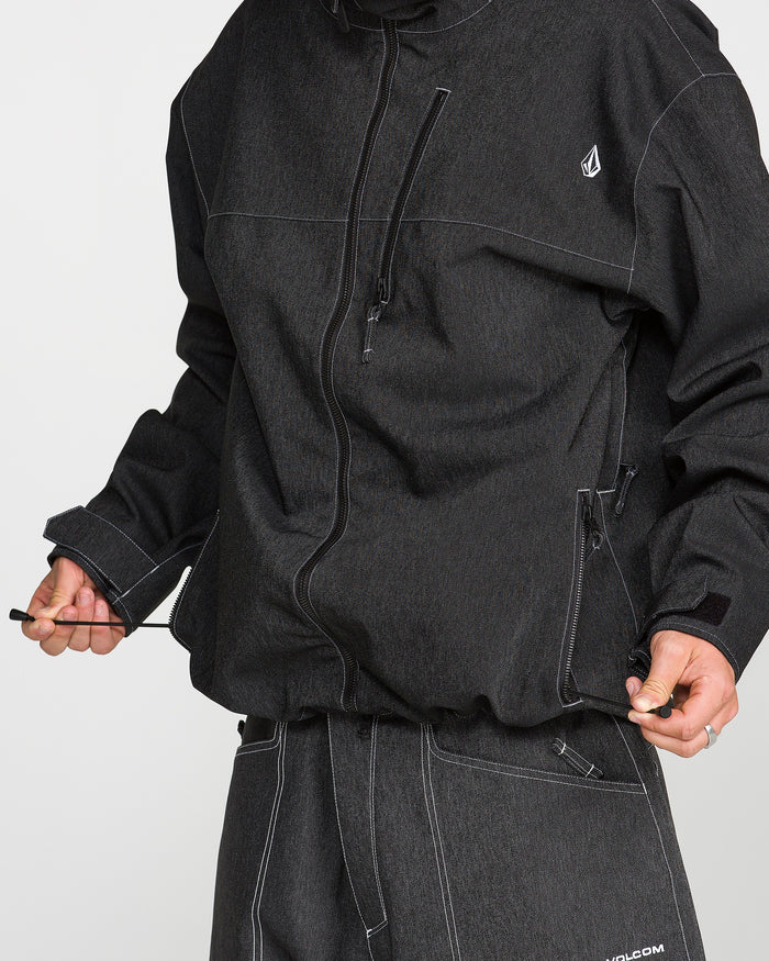 A person is seen wearing the Volcom Men’s Top Stitch Jacket in dark gray with matching pants, holding the jacket’s drawstring at the hem. This baggy fit jacket showcases white stitching and a small logo on the chest. The person’s face is not visible.
