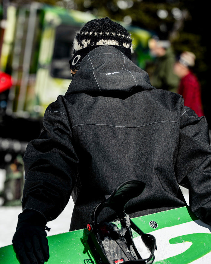 A person, seen from behind, is wearing the Volcom Men’s Top Stitch Jacket in black with a baggy fit and a patterned hat, holding a green snowboard outdoors. Blurred people and a green vehicle appear in the background. The 10K waterproof Volcom jacket features a PFAS-free DWR finish for enhanced protection and sustainability.
