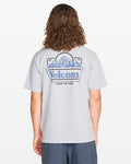 A person with long, curly hair wears a light gray Volcom Men's Topas Short Sleeve Tee featuring a large blue mountain and Volcom logo graphic, with “TRUE TO THIS” printed below on the back.