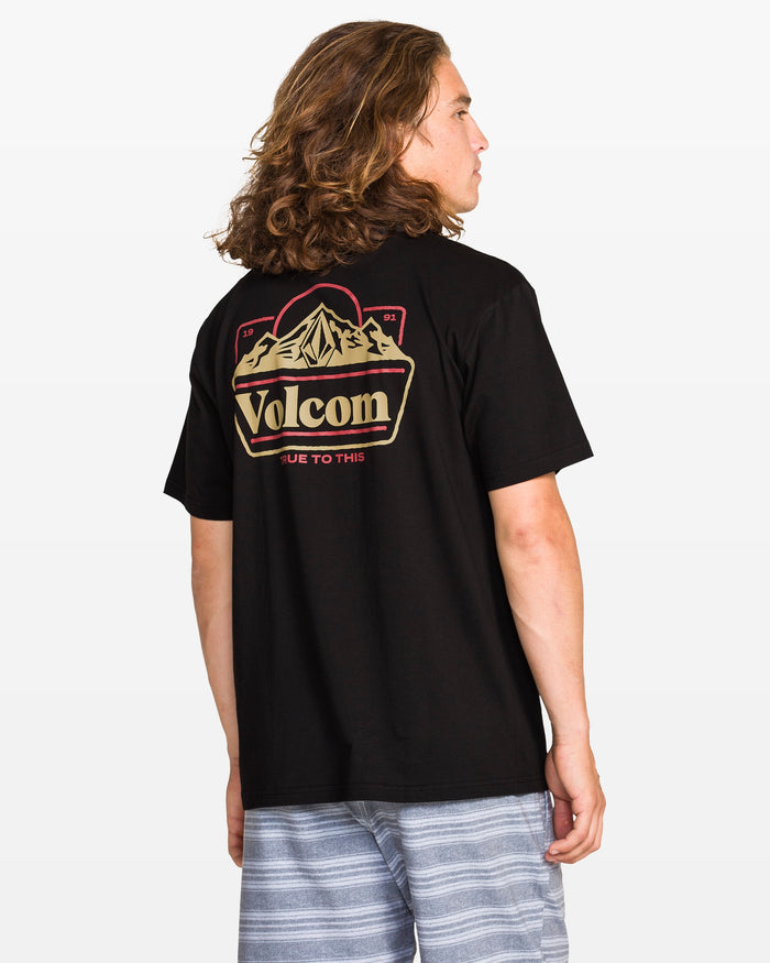 A person with long hair faces away, wearing Volcom's Men's Topas Short Sleeve Tee in black with a mountain graphic on the back, paired with light gray striped shorts. The background is plain white.