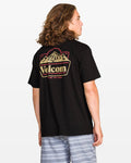 A person with long hair faces away, wearing Volcom's Men's Topas Short Sleeve Tee in black with a mountain graphic on the back, paired with light gray striped shorts. The background is plain white.