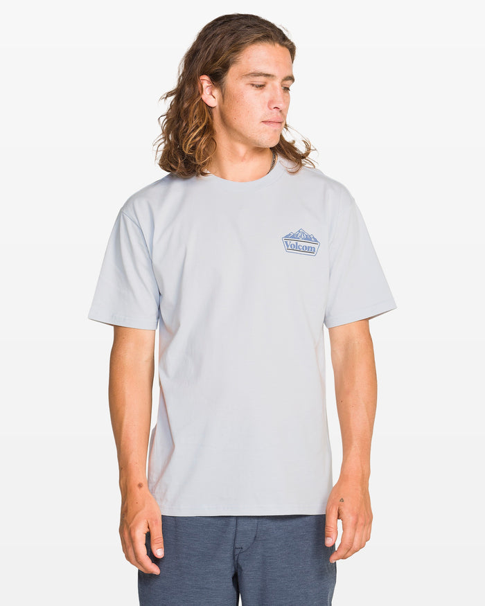 A man with long brown hair wears the Volcom Men's Topas Short Sleeve Tee in light grey and dark shorts, standing against a plain white background while looking down and to his left.