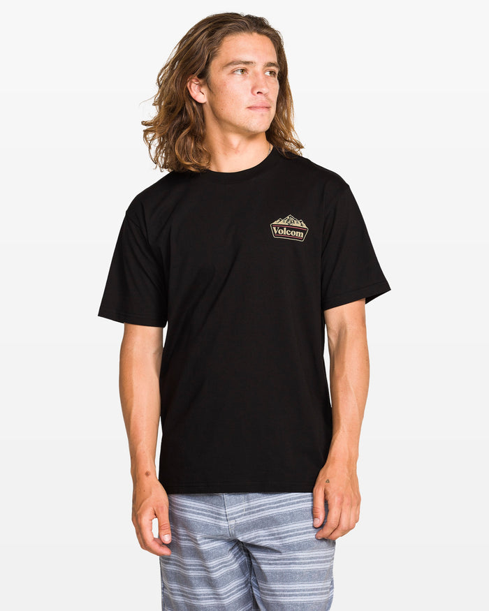 A person with long hair wears a black Volcom Men's Topas Short Sleeve Tee and light blue striped shorts, standing against a plain white background and looking slightly to the side.