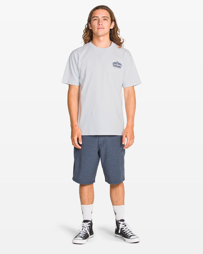A person with long brown hair stands facing forward, wearing the Volcom Men's Topas Short Sleeve Tee, navy blue shorts, white crew socks, and black high-top sneakers against a plain white background.