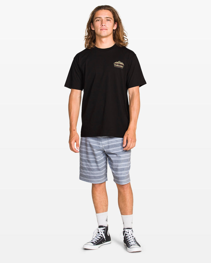 A young man with long hair stands facing forward, wearing the Volcom Men's Topas Short Sleeve Tee in black, gray striped shorts, white socks, and black high-top sneakers against a plain white background.