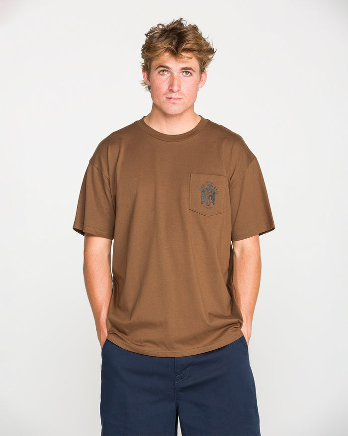 A person with short, tousled hair wears the Volcom Men's Troubador Short Sleeve Tee—a brown, loose-fit shirt made from heavyweight ringspun cotton with a small chest pocket—and pairs it with navy blue shorts while standing against a plain white background.