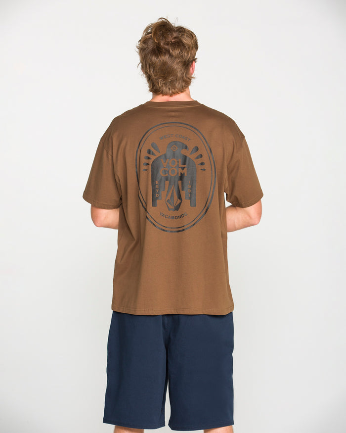 A person with short, light brown hair is standing facing away, wearing the Volcom Men's Troubador Short Sleeve Tee—a brown heavyweight ringspun cotton T-shirt featuring a large circular graphic on the back—paired with navy blue shorts, against a plain white background.