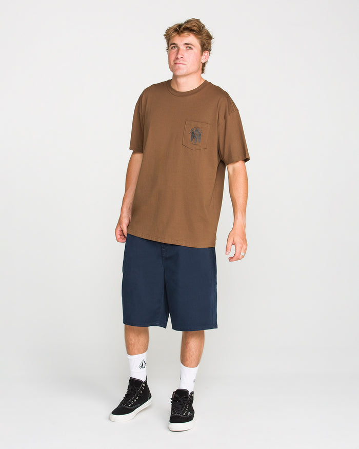 A young man stands against a plain white background wearing Volcom’s Men's Troubador Short Sleeve Tee in brown, crafted from heavyweight ringspun cotton with a practical chest pocket, paired with navy blue shorts, white socks, and black sneakers for a laid-back look.