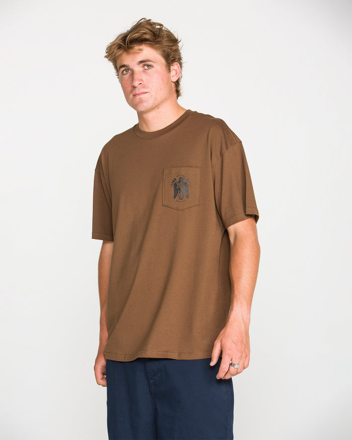 A young man with light brown hair poses against a white background, wearing the Volcom Men's Troubador Short Sleeve Tee—a loose-fitting, heavyweight ringspun cotton t-shirt in brown featuring a chest pocket embellished with a small black graphic. He pairs it effortlessly with dark navy trousers.