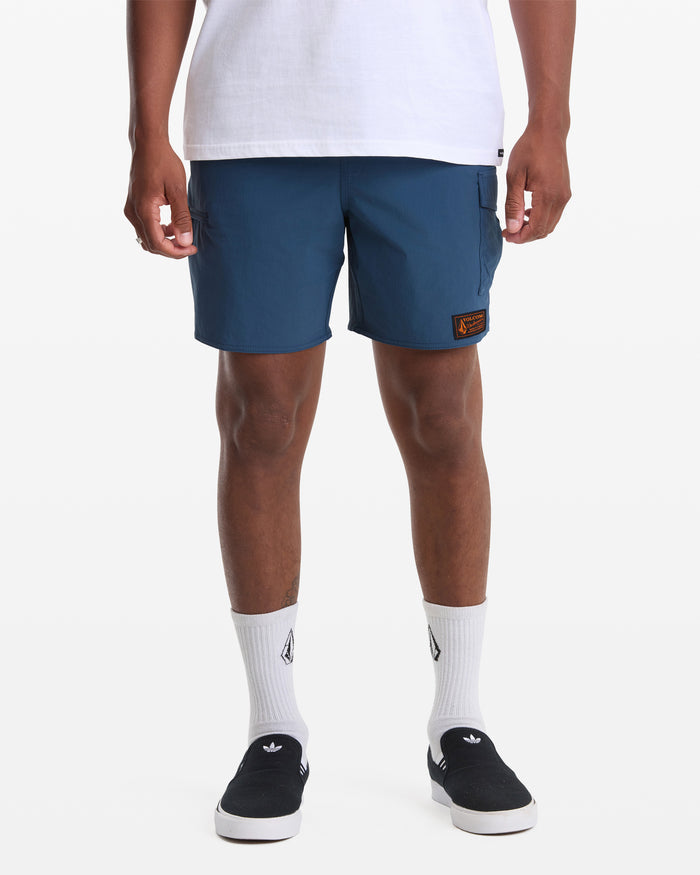 A person in a white T-shirt, Volcom Men's Truly Lib Work Trunk 17 blue shorts with a patch on the left leg, white logo socks, and black slip-on shoes stands against a plain white background.