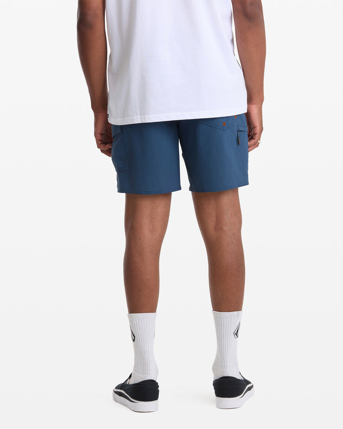 A man wearing a white shirt, white socks, black slip-on shoes, and Volcom's Men's Truly Lib Work Trunk 17 shorts stands with his back to the camera against a plain white background.