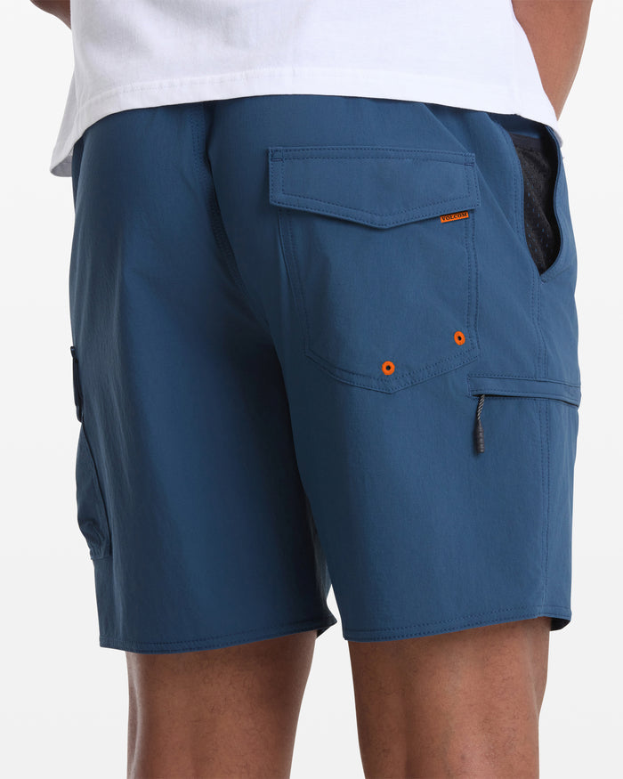 A person wearing Volcom Men's Truly Lib Work Trunk 17 blue cargo shorts with flap pockets and orange accents stands with their back to the camera, showing the lower part of a white shirt.