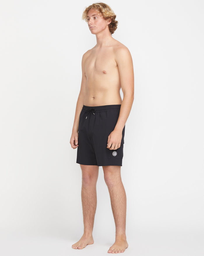 A young man with light skin and blonde hair stands barefoot against a white background, wearing Volcom Men's Truly Liberators 17" black trunk shorts with a circular logo and left-leg cargo pockets. He is shirtless and looking slightly to the side.