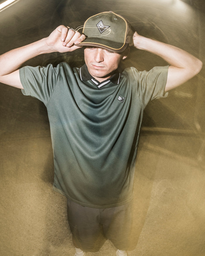 A person in a green shirt and hat stands outdoors at night, holding the brim of their Volcom Men’s V Line Adjustable Hat with both hands. The lighting creates a dreamy, blurred effect around them.