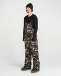A person wearing a black beanie, long-sleeve black shirt, and the Volcom Men’s V.Co Sparta Bib—baggy camo overalls made from recycled fabric—stands against a plain white background, looking slightly to the side.