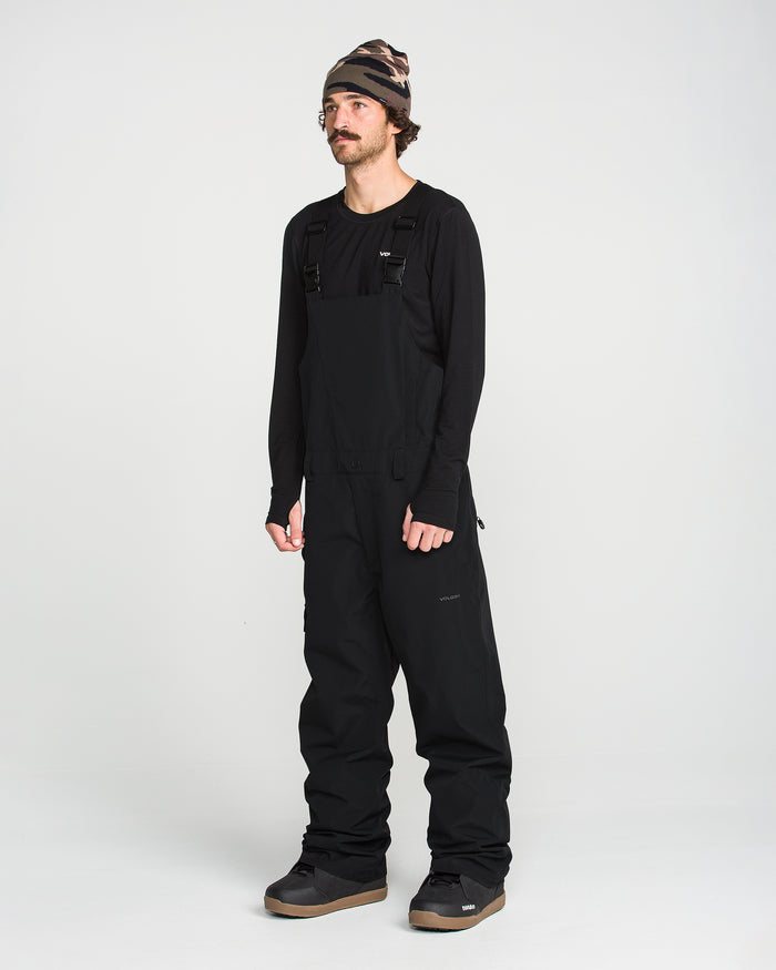 A person wearing a black beanie, a black long-sleeve shirt, and the Volcom Men’s V.Co Sparta Bib—crafted from recycled fabric with waterproof protection—stands against a plain white background.