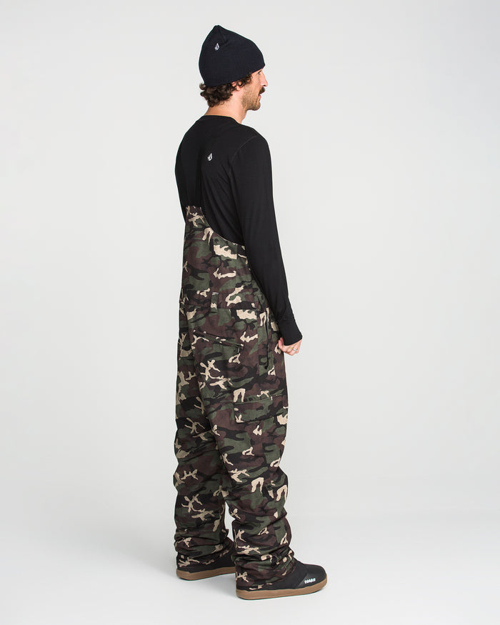 A person stands sideways on a white background wearing the Volcom Men’s V.Co Sparta Bib—camouflage, baggy overalls made from recycled fabric—paired with a black long-sleeve shirt and black beanie.