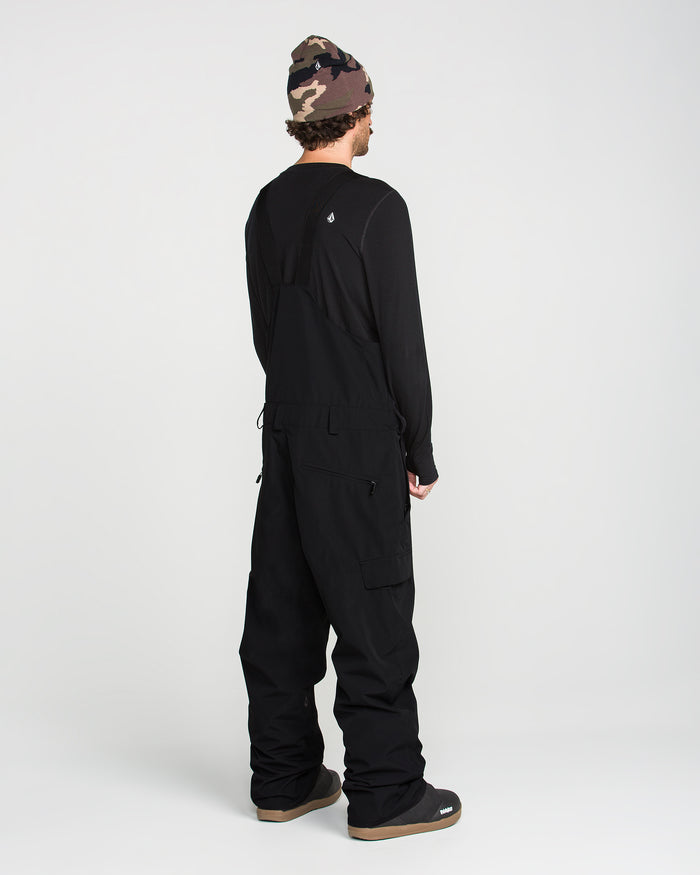 Wearing the Volcom Men’s V.Co Sparta Bib crafted from recycled fabric, a person stands facing away against a plain white background, paired with a black long-sleeve shirt, black boots, and a camouflage beanie.
