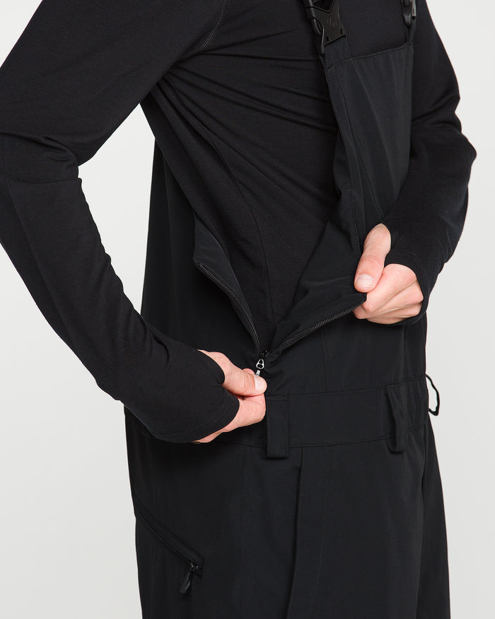 A person dressed in black is unzipping the side zipper pocket of the Volcom Men’s V.Co Sparta Bib, showcasing its waterproof protection; only their torso and hands are visible.