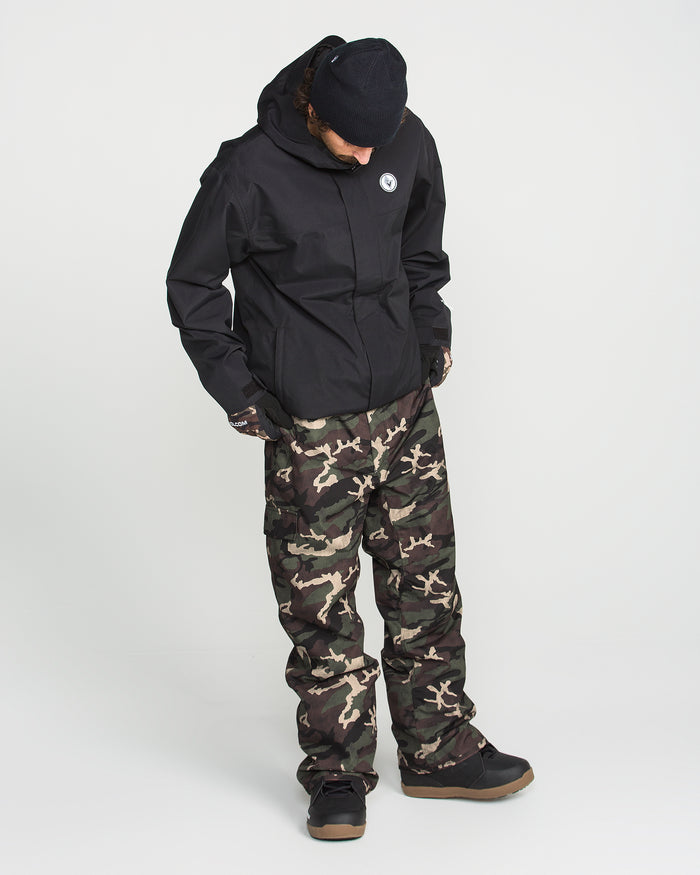 A person stands with hands in pockets against a plain white background, wearing the Volcom Men’s V.Co Sparta Bib, a black hooded jacket with waterproof protection, a black beanie, and camouflage pants.